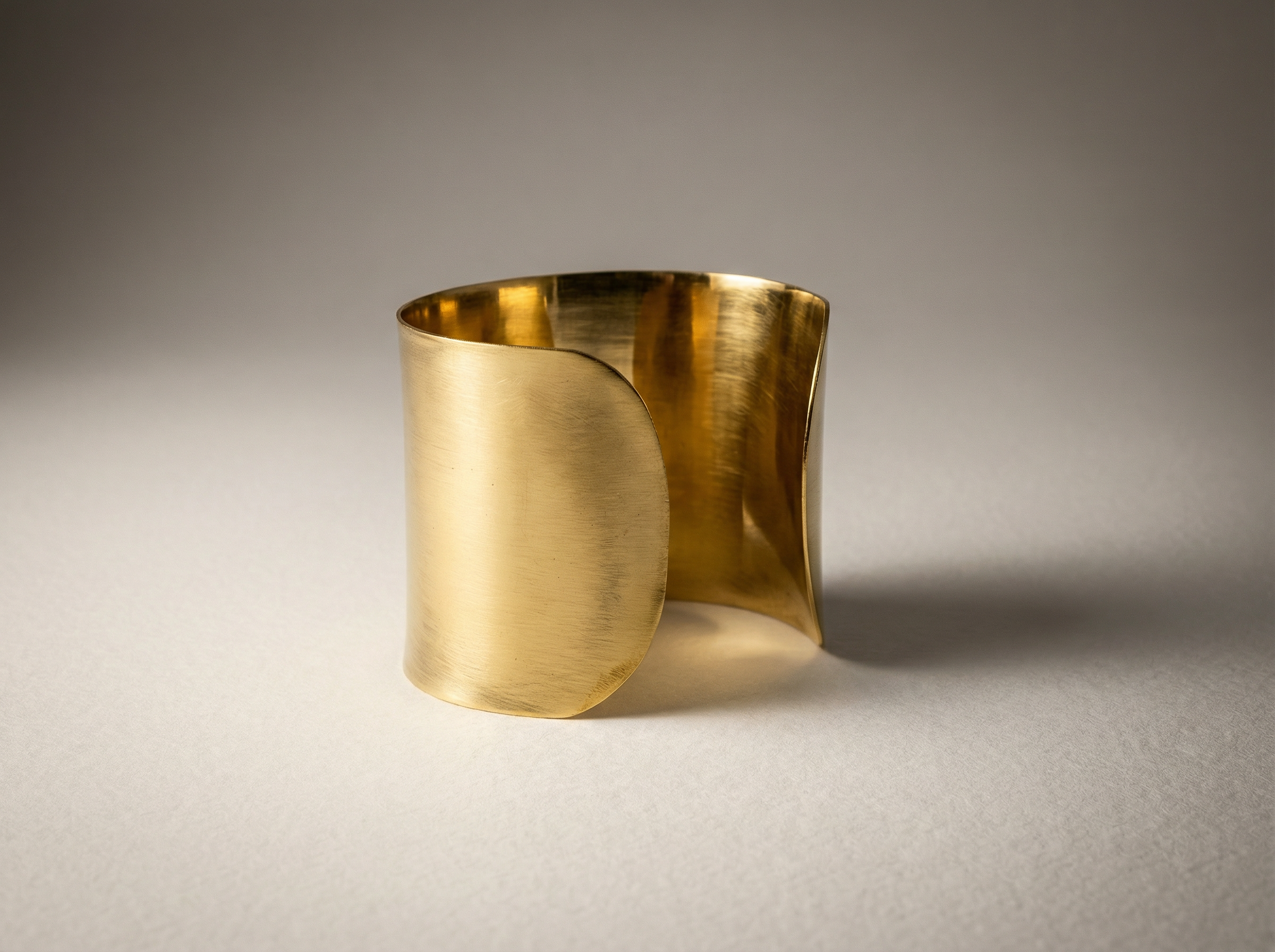 Studio close-up of the Dune Cuff, a wide sculptural gold bracelet with softly curved overlapping edges, standing upright on a light textured surface with a gentle gradient backdrop.