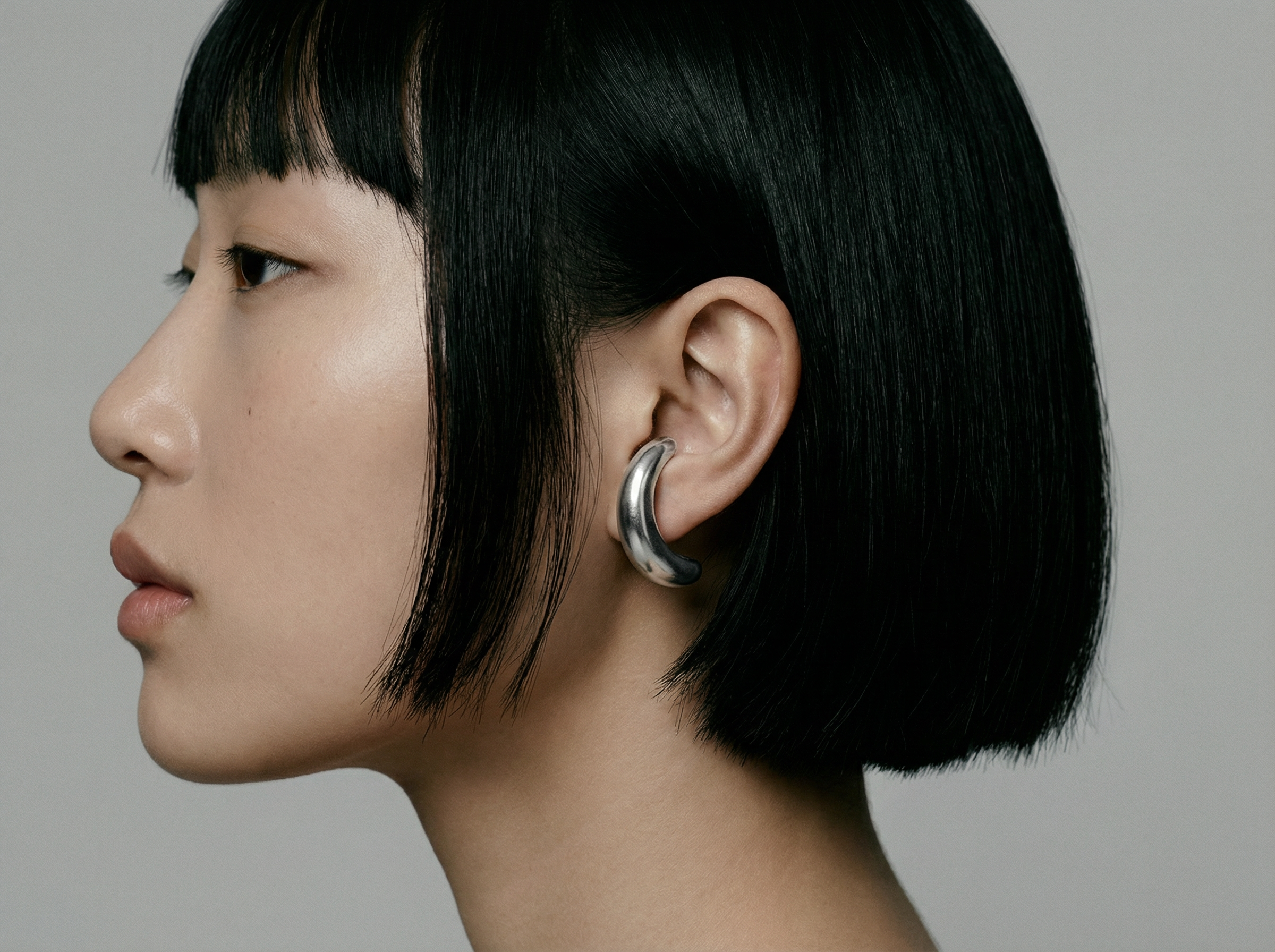 Side view of a woman with a straight black bob wearing Luna Loops, thick crescent-shaped silver hoop earrings that hug the earlobe, against a soft grey background.