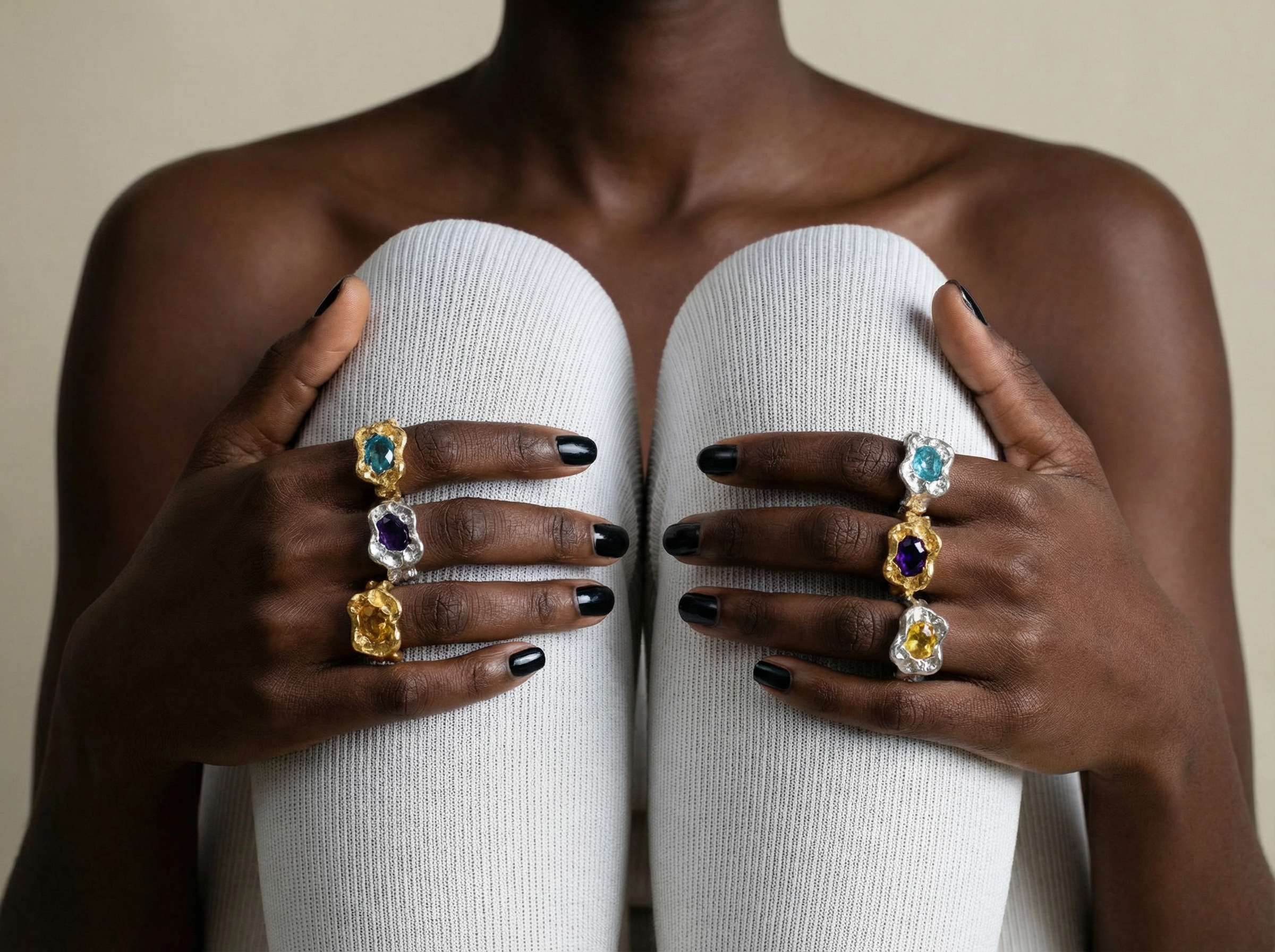 Close-up of a seated model with dark skin wearing six chunky gemstone cocktail rings on both hands, resting on light ribbed knit-covered knees, with black nail polish and a neutral background.