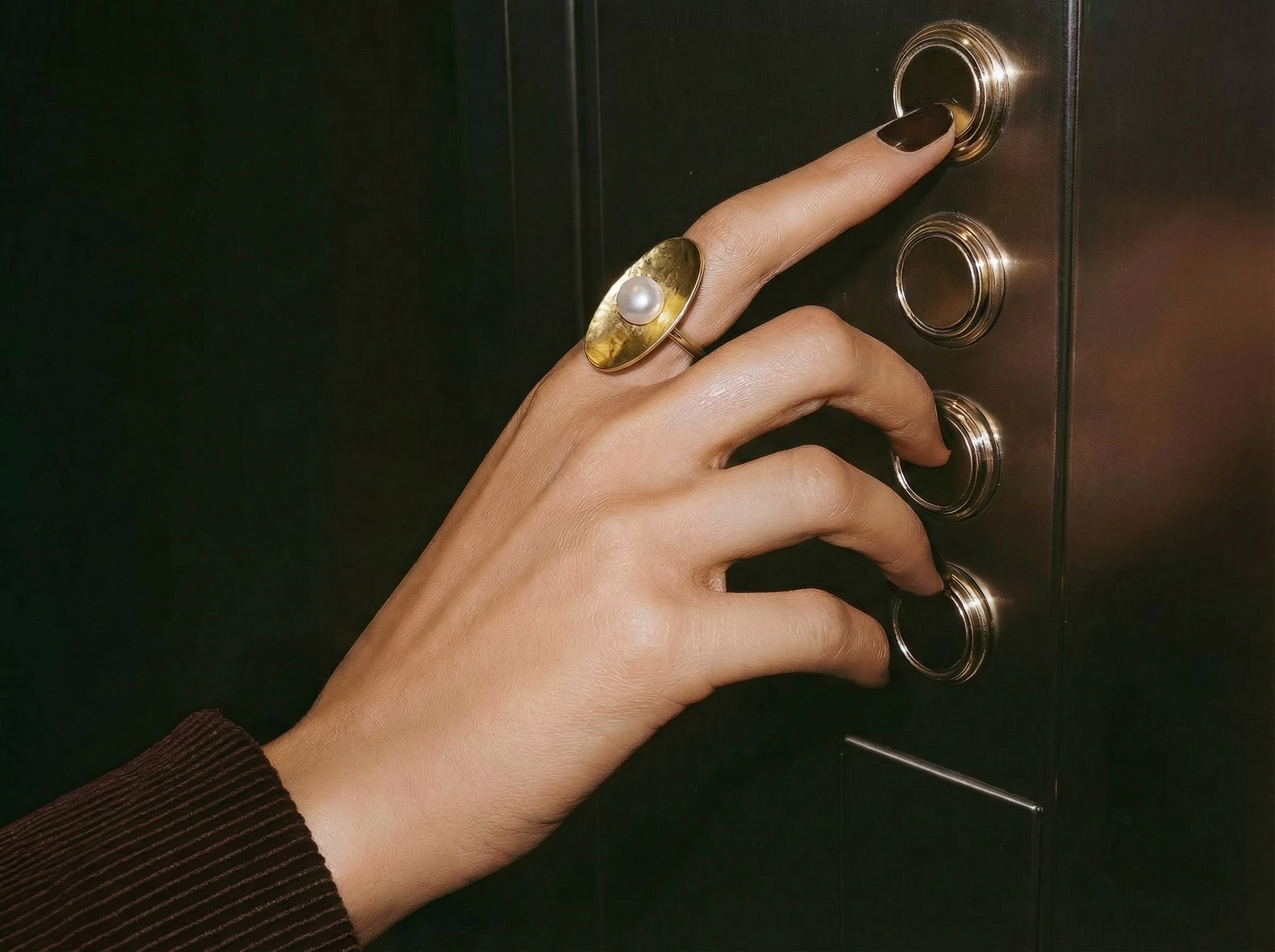Close-up of a hand wearing the Solar Pearl Ring, a large hammered gold dome ring set with a central white pearl, as the model presses an elevator button against a dark metallic control panel.
