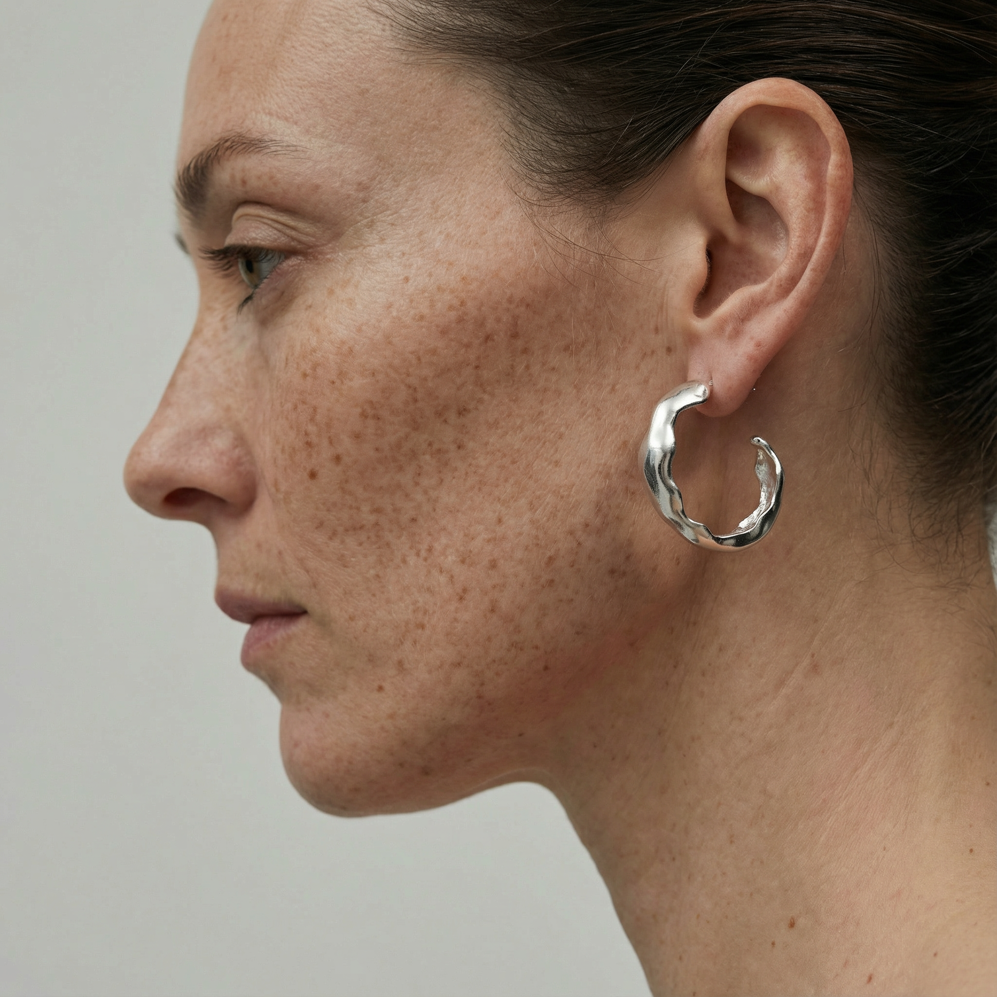 Side profile of a woman with freckles and pulled-back hair wearing the Tirtik Earrings, chunky sculptural silver hoops with an organic wavy edge, against a soft grey background.