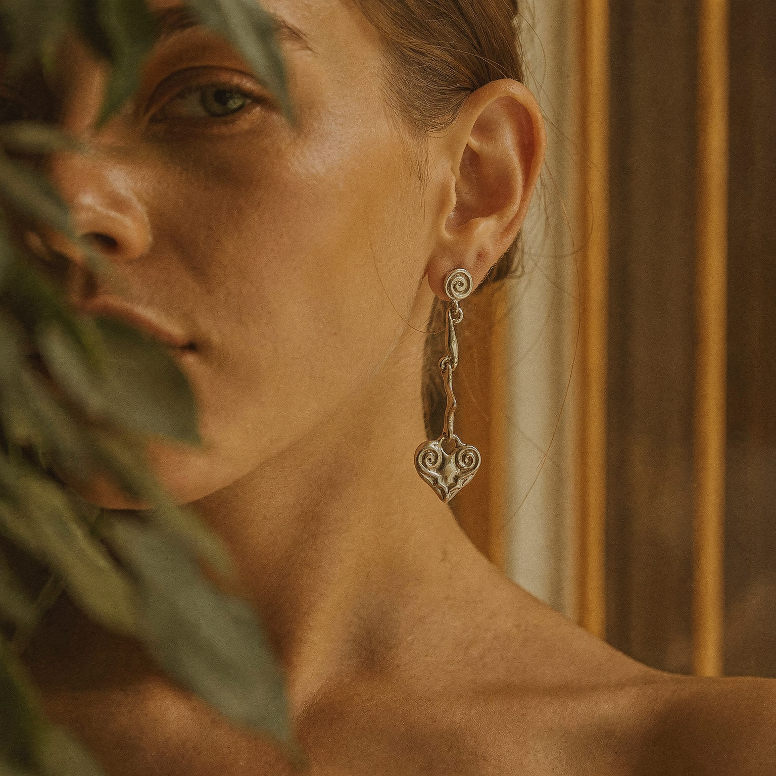 Close-up of a woman’s bare shoulder and jawline wearing a long artisan silver drop earring with a scroll heart pendant, partially framed by soft-focus green leaves and warm wooden background.