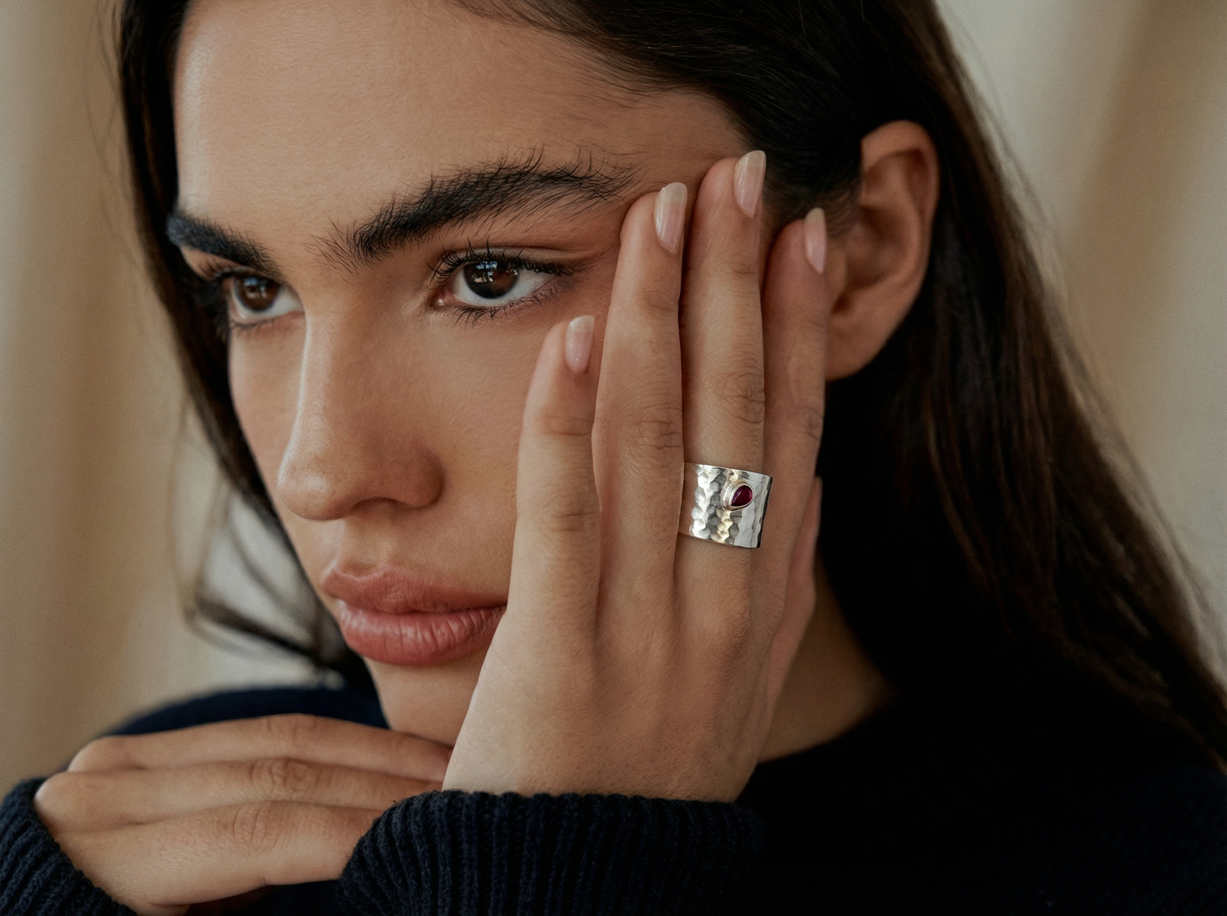 Close-up of a woman resting her face on her hands, wearing a wide hammered silver ring with a small marquise ruby stone, styled with a dark knit sweater and neutral background.