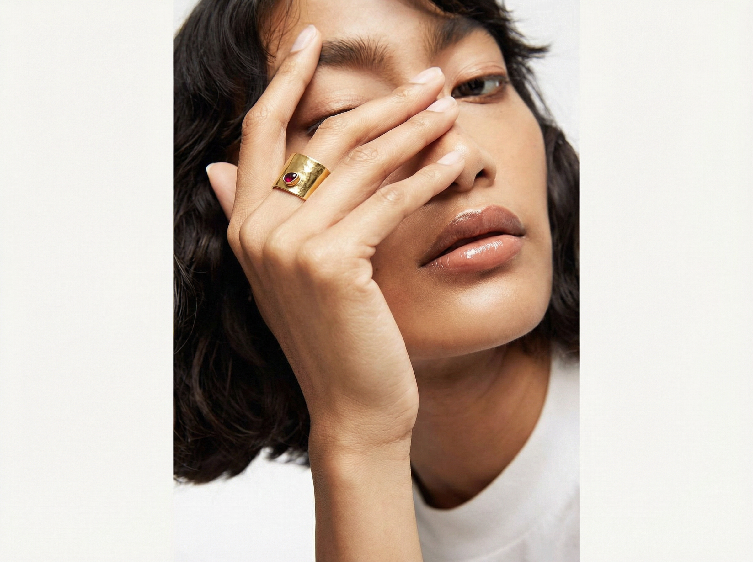 Close-up of a woman covering one eye with her hand while wearing the Pulse Ring, a wide polished gold band set with a single round red gemstone, styled with a white top and clean background.