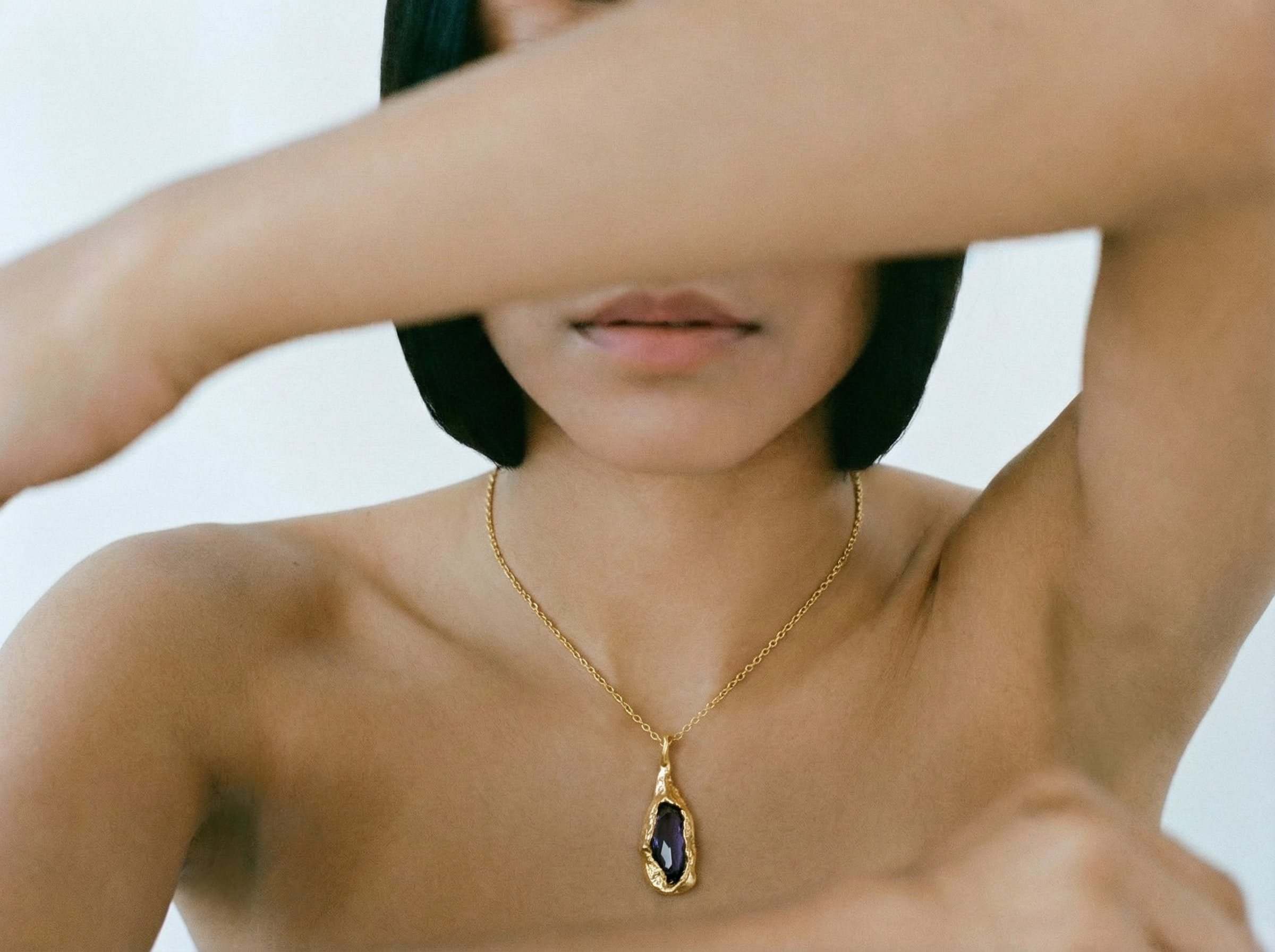 Bare-shouldered model with short dark hair wearing the Magma Pendant, a gold teardrop-shaped setting holding a deep purple stone on a fine gold chain, with her arms raised across the frame against a light background.