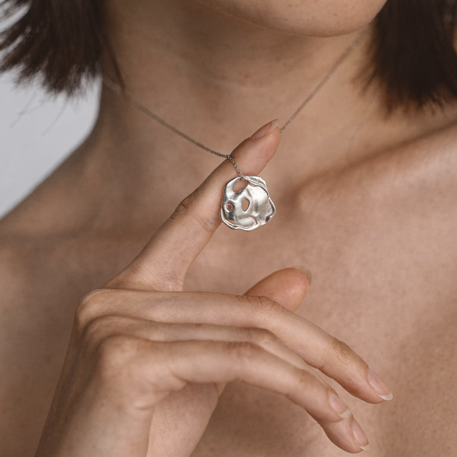 Close-up of a bare neckline wearing the Orogeny necklace, featuring an irregular sculptural silver pendant with organic cutout holes, held delicately between the model’s fingers against a soft neutral background.