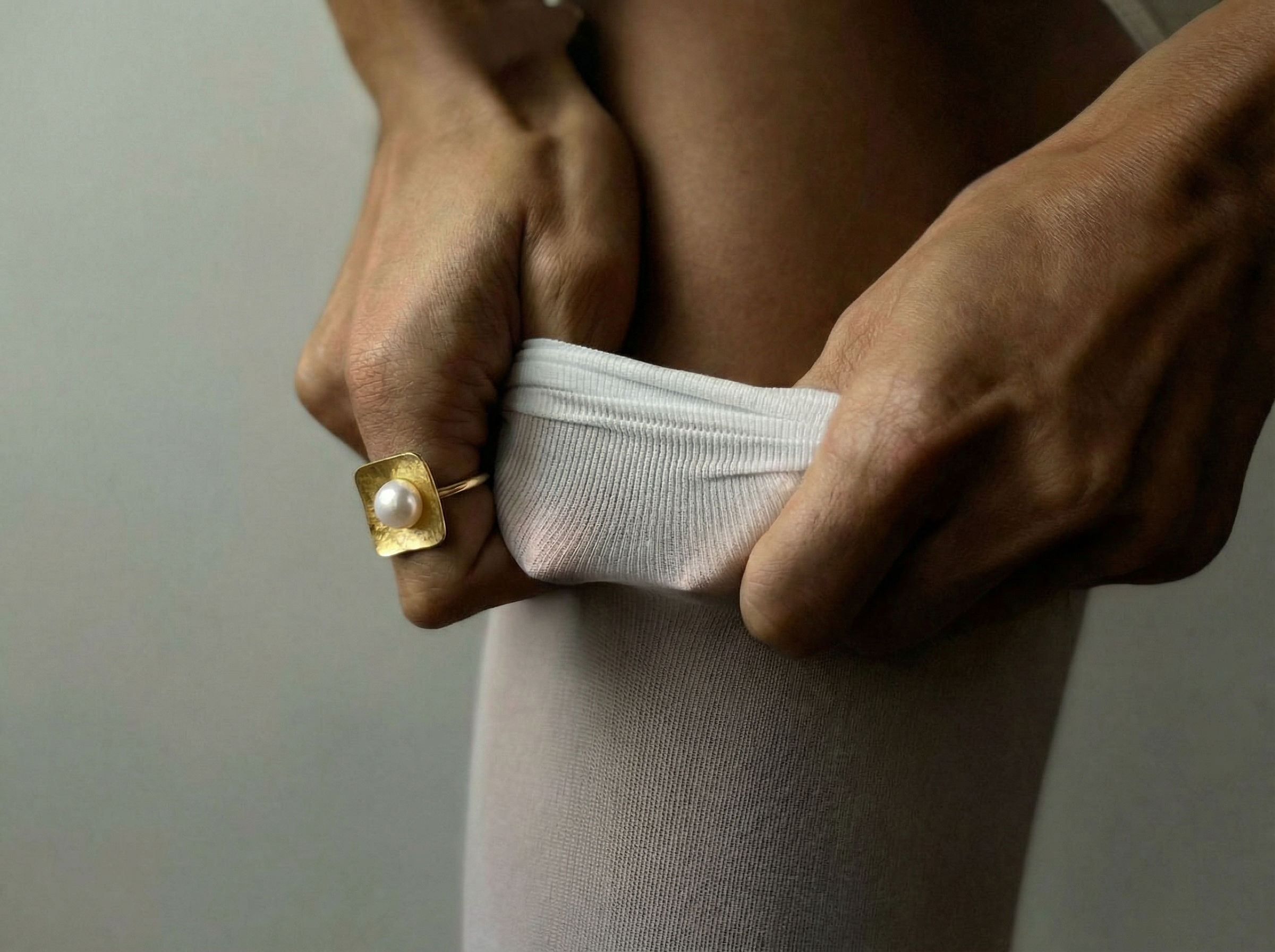 Close-up of hands pulling up soft white ribbed tights while wearing the Shell Light Ring, a square hammered gold ring set with a single round white pearl, against a muted neutral background.